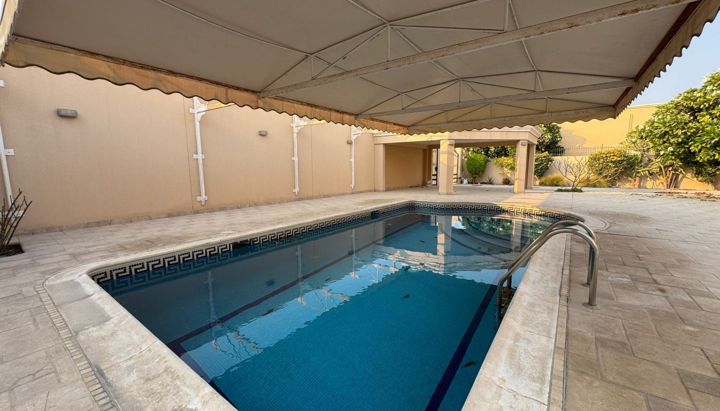 Rectangular outdoor swimming pool with blue tiles, partially covered by a large canopy, surrounded by a tiled patio and cream-colored walls.