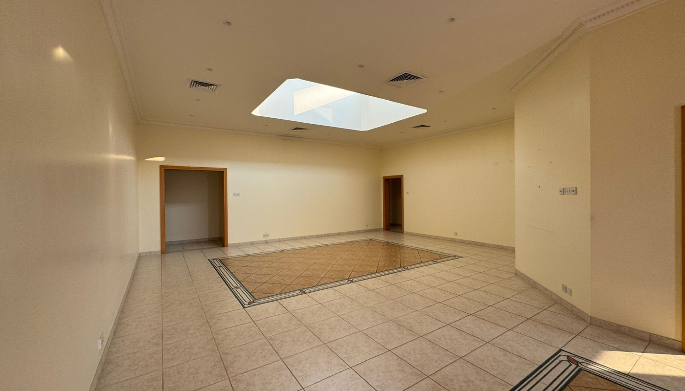 Spacious empty room with tiled floors, cream-colored walls, a central square skylight, and multiple doorways leading to adjacent areas.