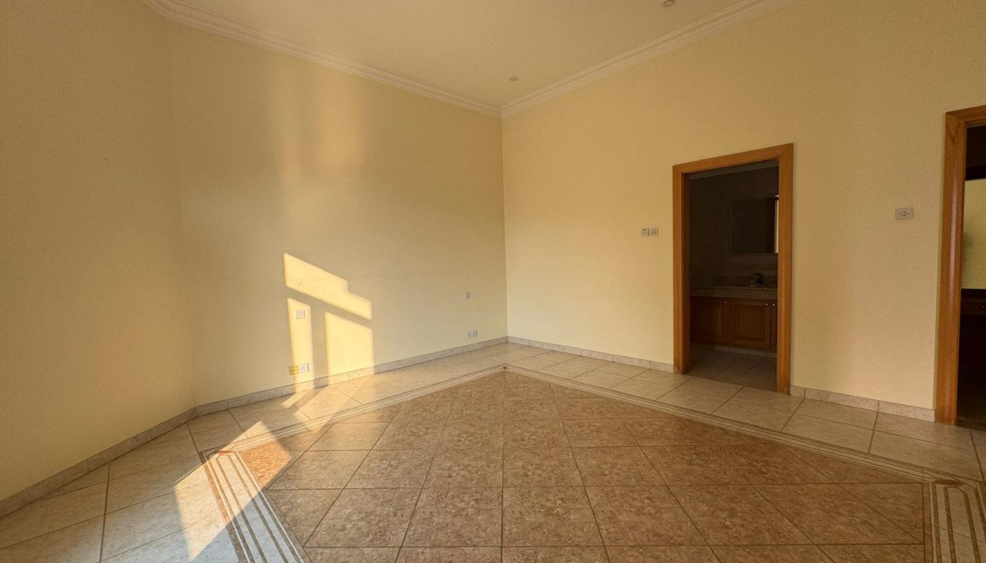 Empty room with beige tiled floor, cream walls, square ceiling vent, and two doorways leading to other rooms; sunlight casts shadows on the wall and floor.