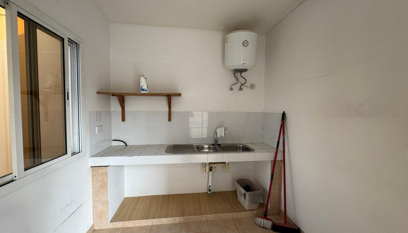 A simple kitchen area with a sink, a shelf holding a bottle, a water heater above, and a broom and dustpan leaning against the wall.