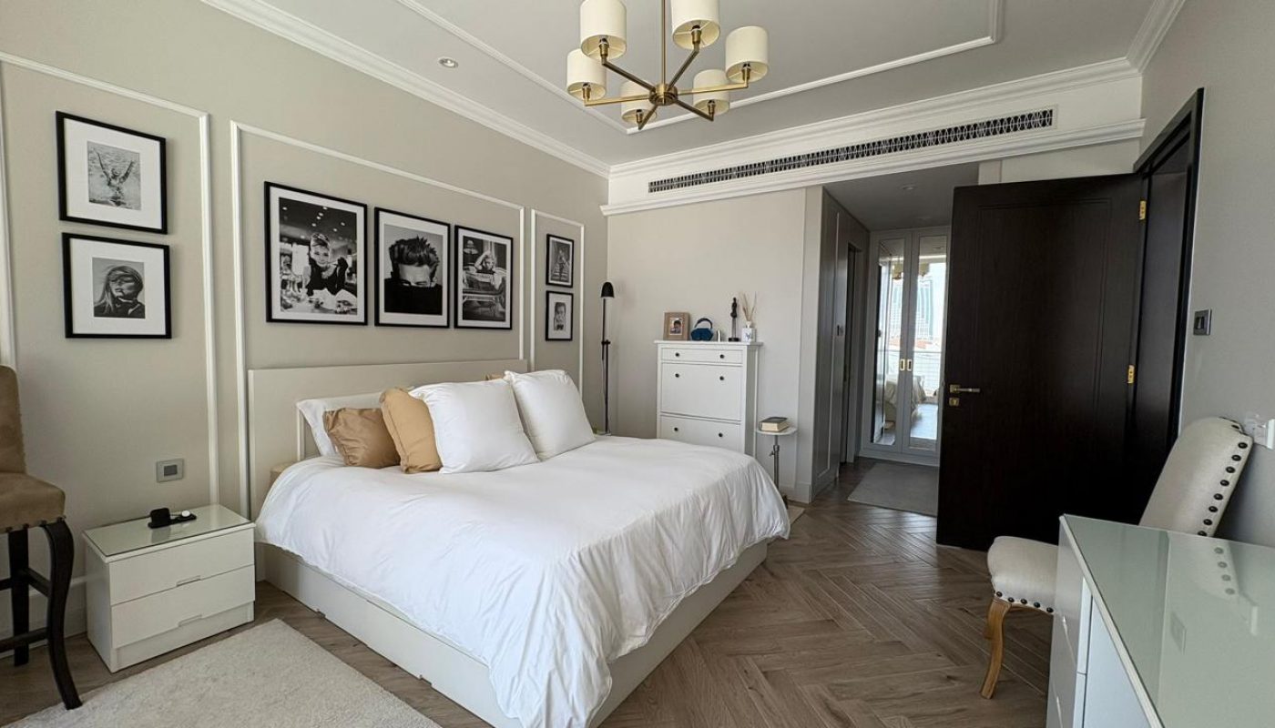A modern bedroom with a white bed, framed photos on the wall, a dresser, a chandelier, and neutral decor. A door leads to an adjacent room with natural light.