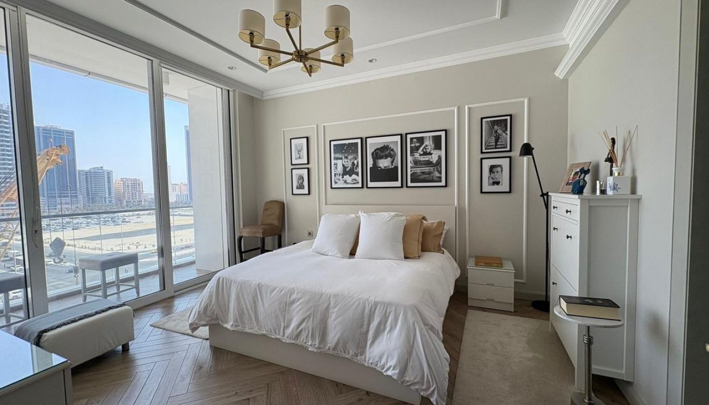 A modern bedroom with a white bed, black and white photos on the wall, large windows showing city buildings, a white dresser, and minimalist decor.