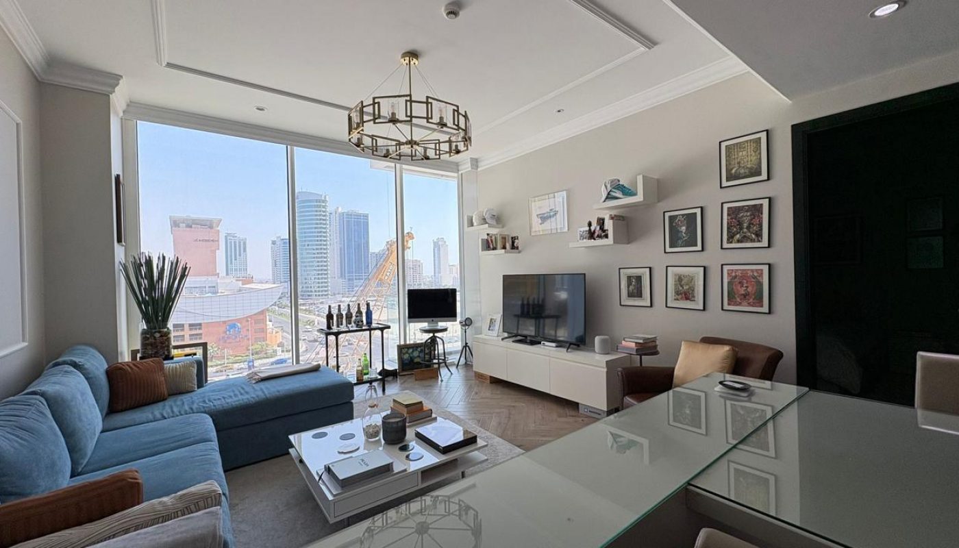 Modern living room with large windows, city skyline view, blue sofa, glass coffee table, TV, wall shelves, and framed art on the wall. Natural light fills the space.