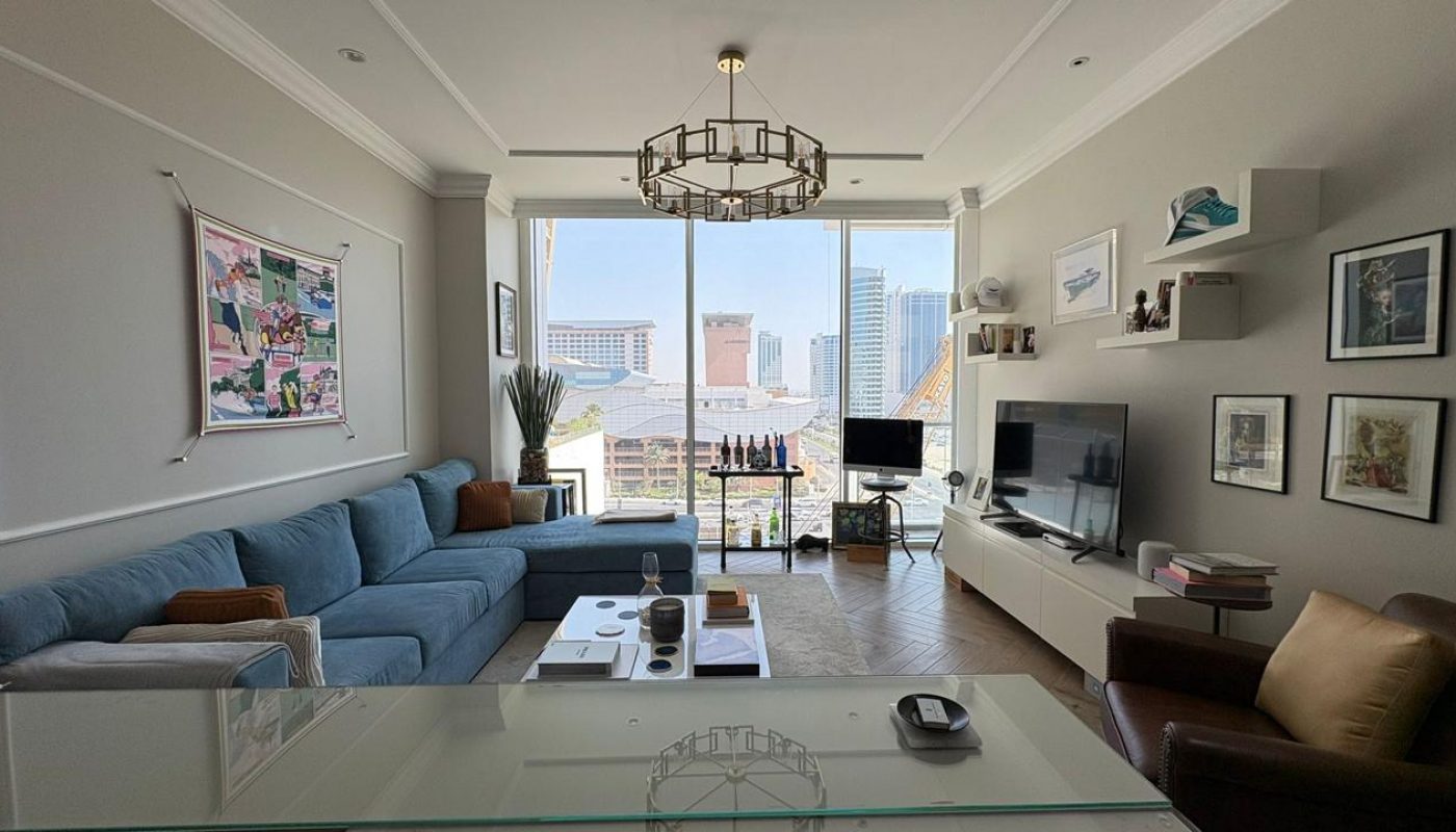 Modern living room with a blue sectional sofa, coffee table, TV, wall art, shelves, and large windows offering a city view with buildings visible outside.