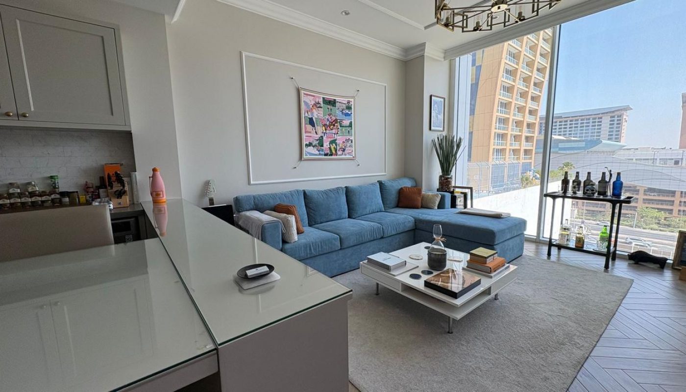 A modern living room with a blue sectional sofa, a glass coffee table with various items, large window, city view, and adjacent kitchen area.