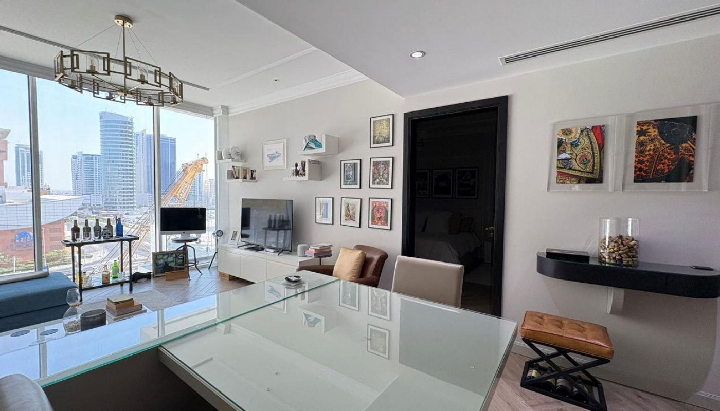 Modern living room with large windows, city view, glass table, wall-mounted TV, art on the walls, and a display of wine bottles by the window.