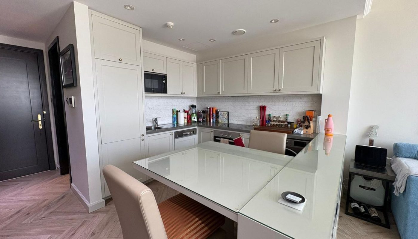 Modern kitchen with light-colored cabinets, built-in appliances, and a glass dining table with beige chairs. Various utensils and kitchen items are on the countertop.