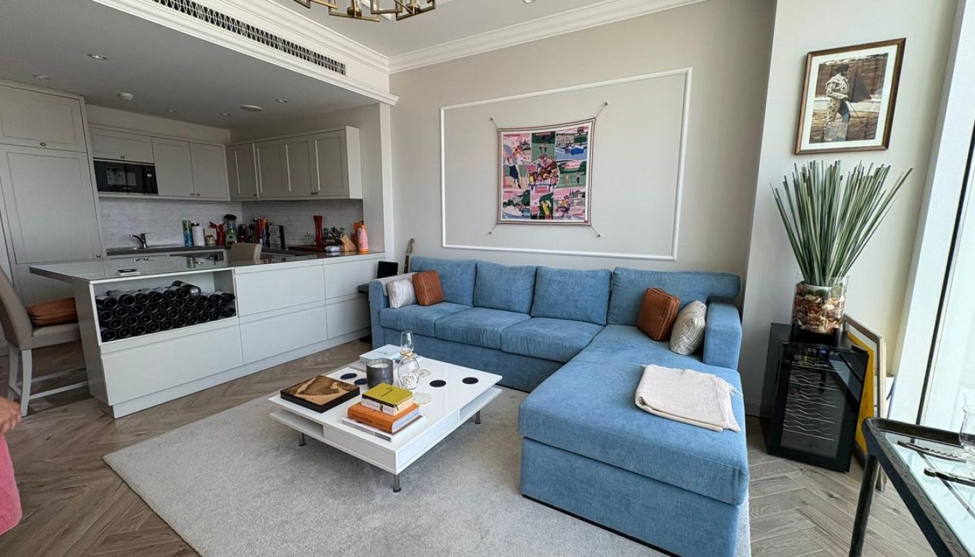 Modern living room with a blue sectional sofa, white coffee table, kitchen area in the background, and artwork on the wall. Natural light streams in from large windows.