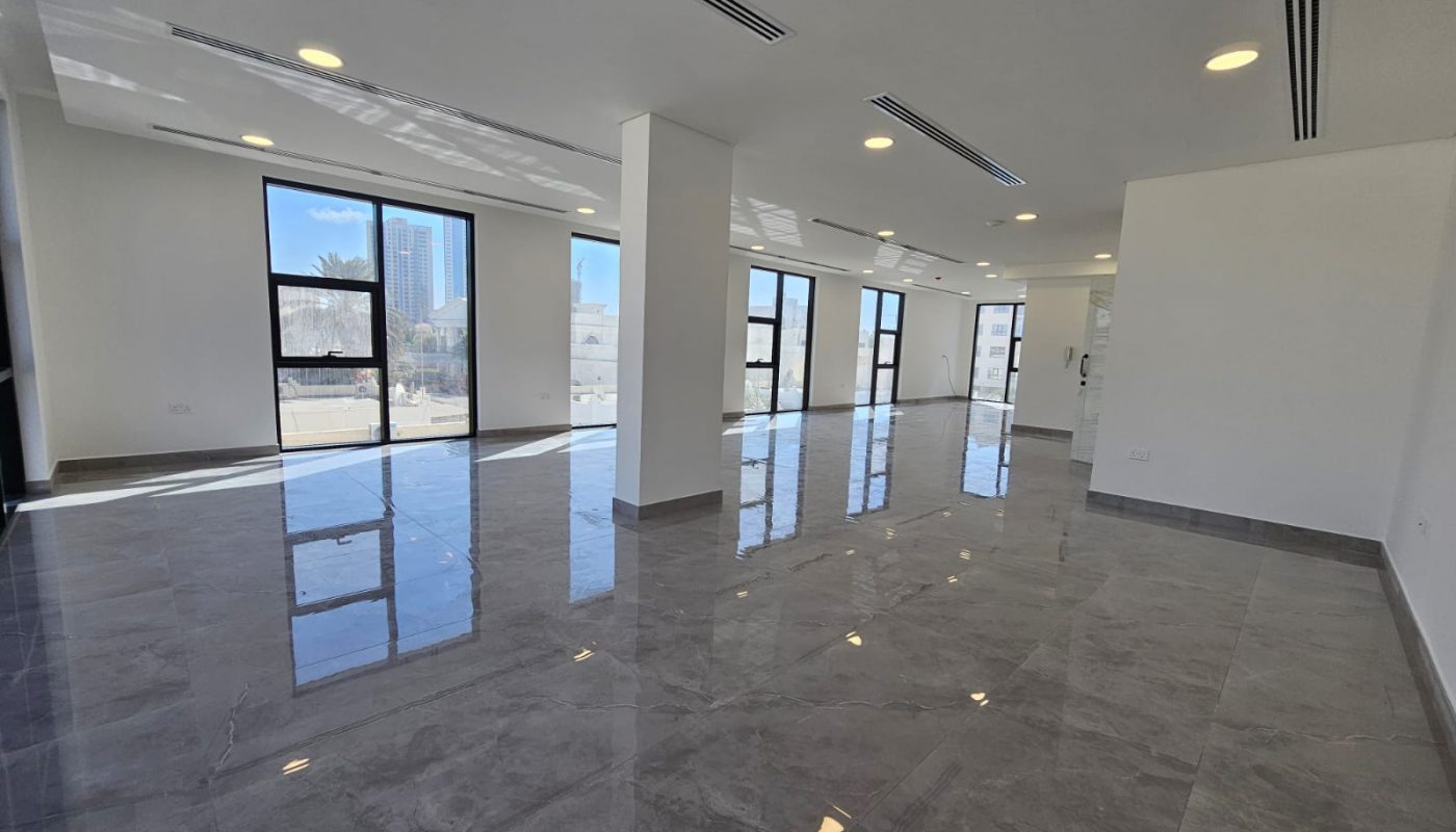 Spacious, empty room with large windows, gray tiled floor, white walls, and ceiling lights. Natural light shines through the windows, reflecting on the floor.