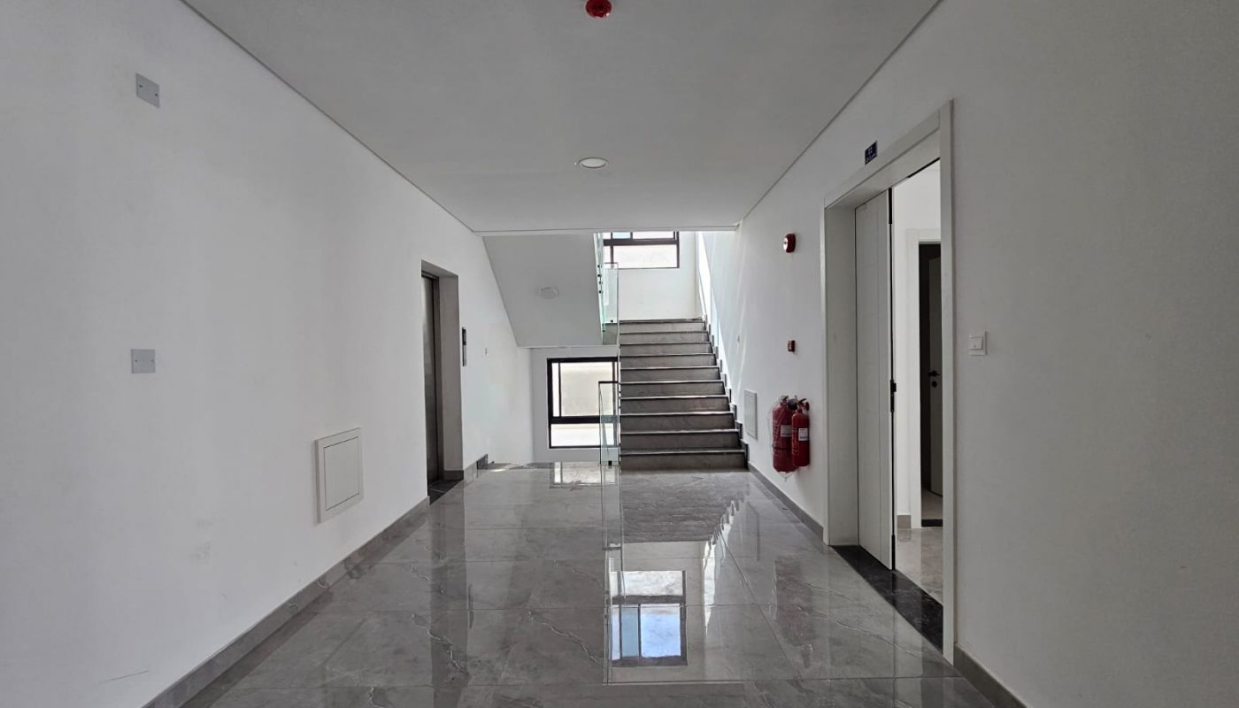 A clean, empty hallway with gray marble floors, white walls, doors on the right, fire extinguishers mounted on the wall, and a staircase leading to an upper floor.