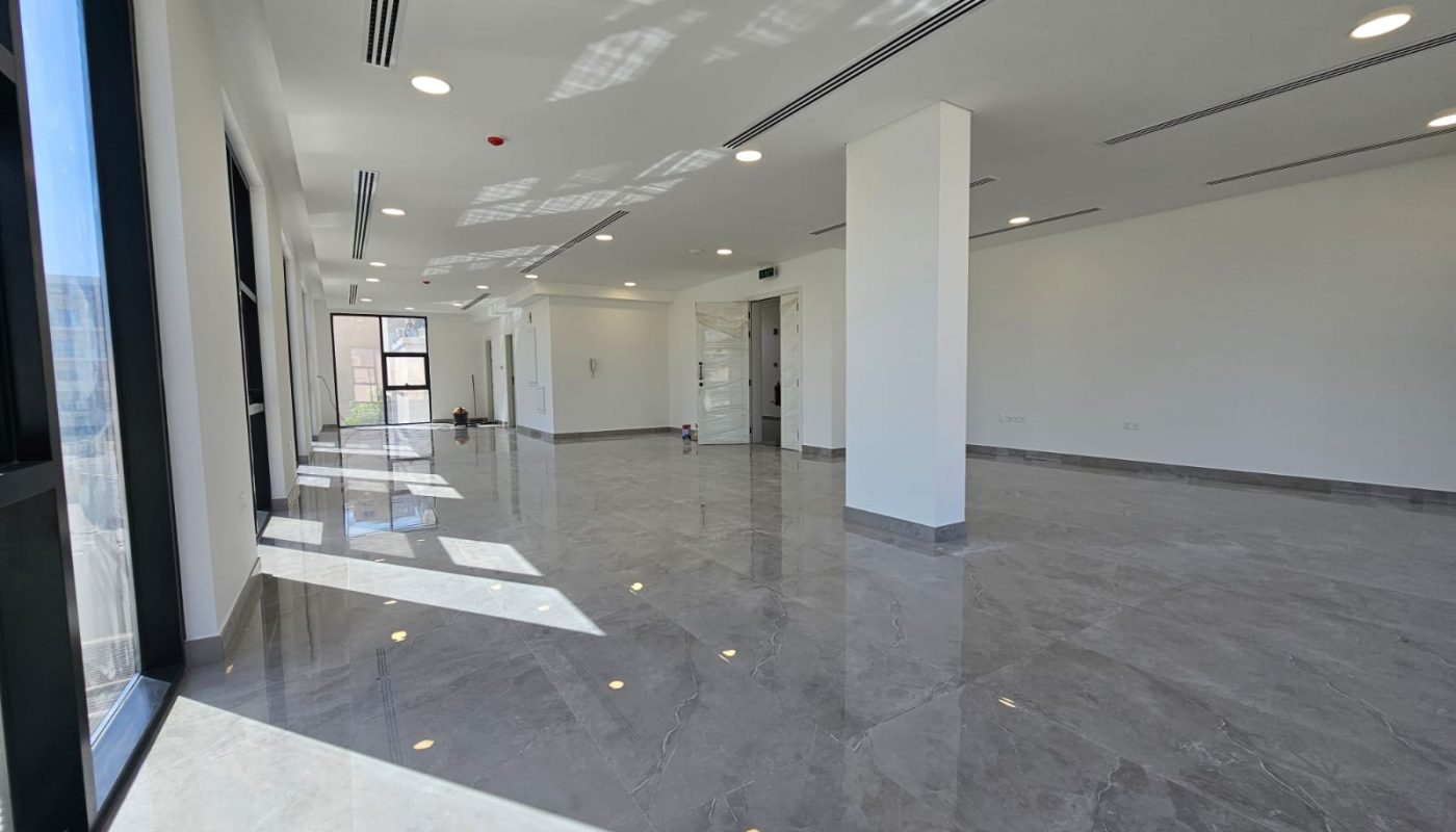 Spacious, empty room with white walls, large windows, shiny gray marble floor, ceiling lights, and square support columns. Bright daylight streams in through the windows.
