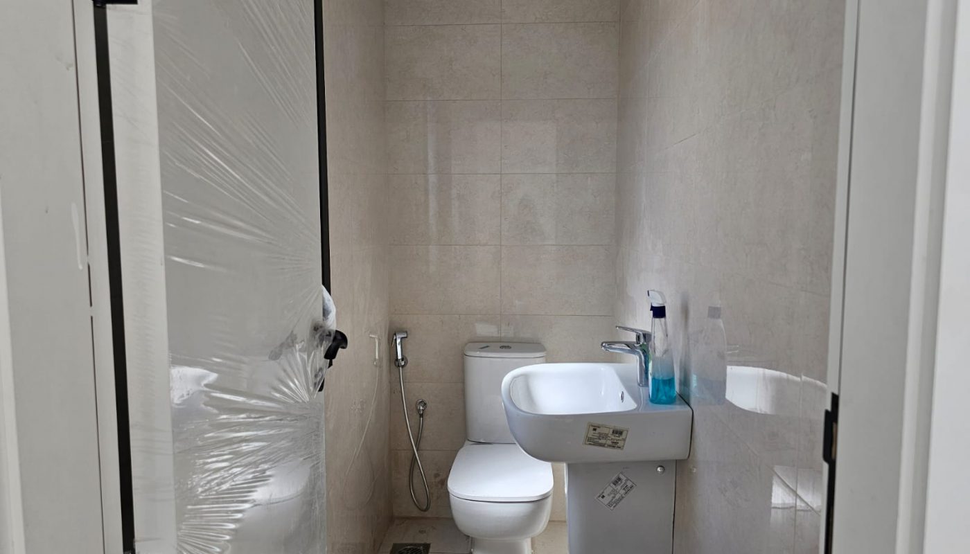 A small bathroom with a toilet, bidet spray, wall-mounted sink, soap dispenser, and cleaning bottles; door partially open and covered in plastic.
