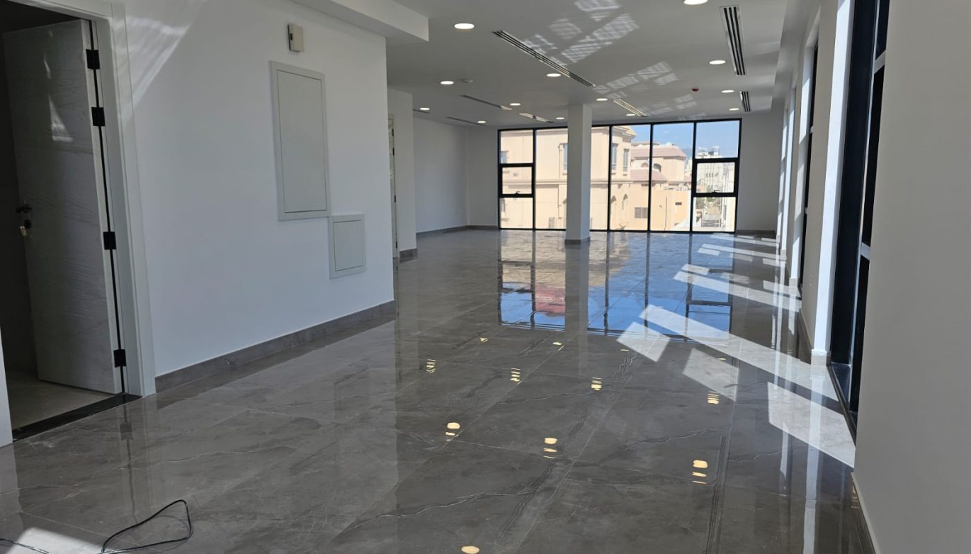 Spacious, empty room with large windows, glossy marble floor, white walls, ceiling lights, and an open door leading to another area. Sunlight streams through the windows.
