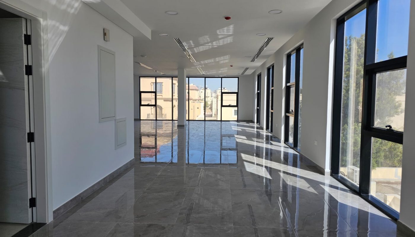 A bright, empty hallway with large floor-to-ceiling windows on the right, glossy tile flooring, and white walls reflecting natural light.