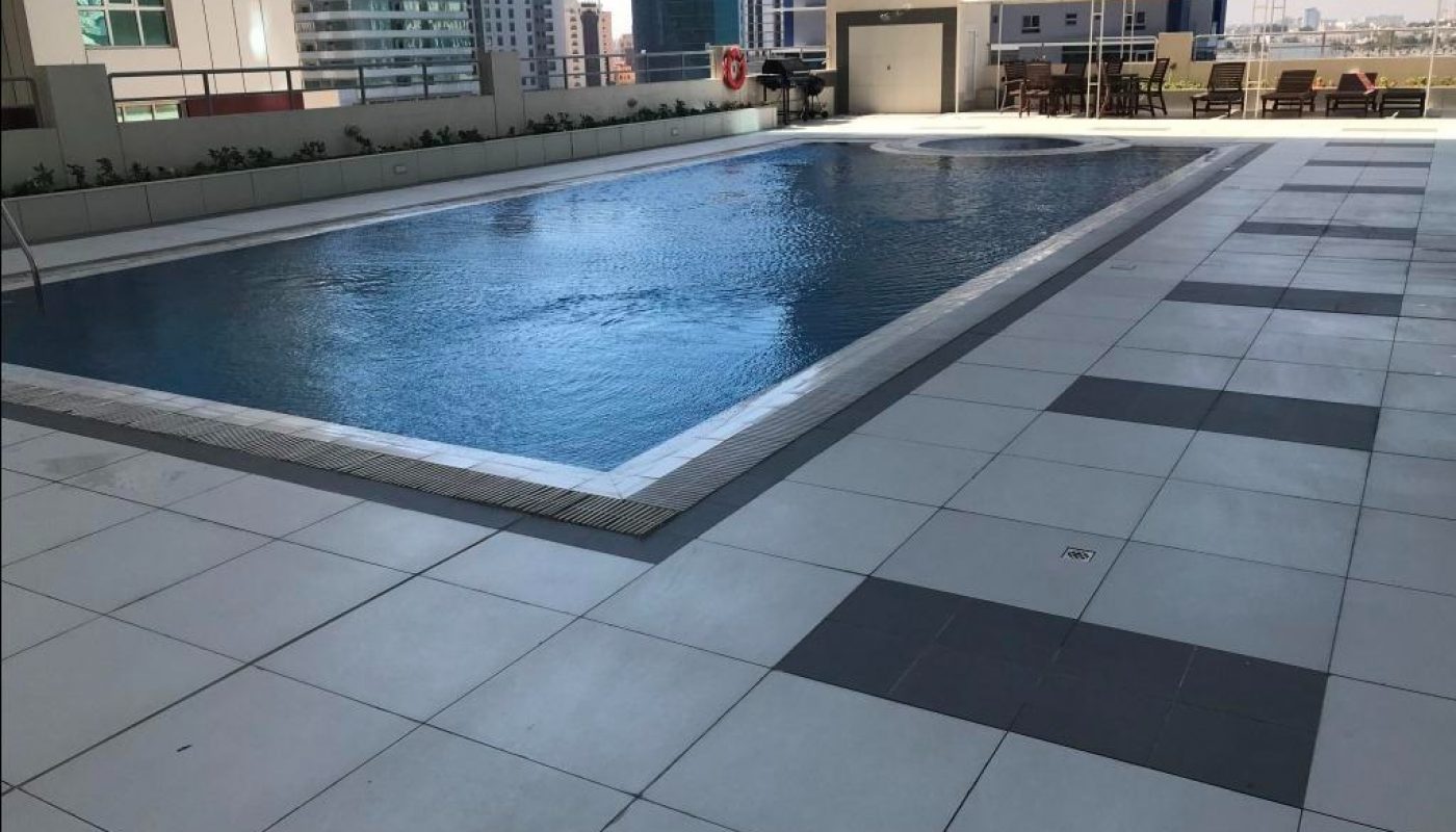 Rooftop swimming pool with surrounding tiled deck, lounge chairs, and umbrellas, set against a backdrop of modern city buildings.