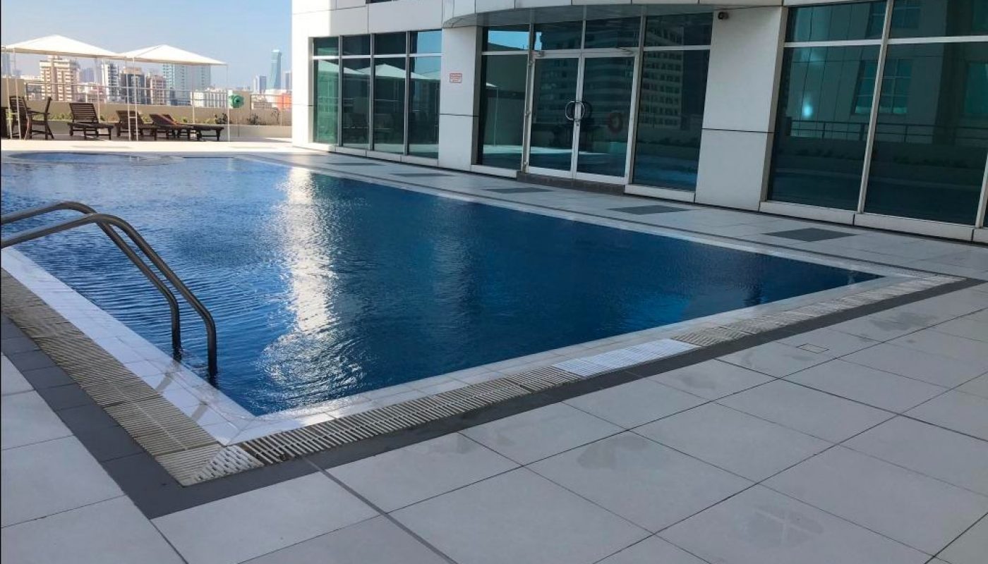 Outdoor swimming pool on a tiled deck beside a modern glass building, with lounge chairs and umbrellas visible in the background.