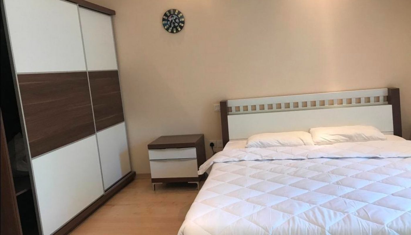 A neatly made bed with white bedding, a wooden headboard, a matching nightstand, a wardrobe with sliding doors, and a wall clock above.