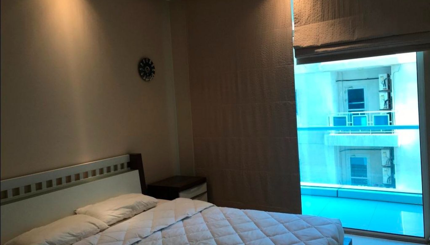 A bedroom with a double bed, two pillows, a nightstand, wall clock, and large window covered by blinds, overlooking a balcony and adjacent building.