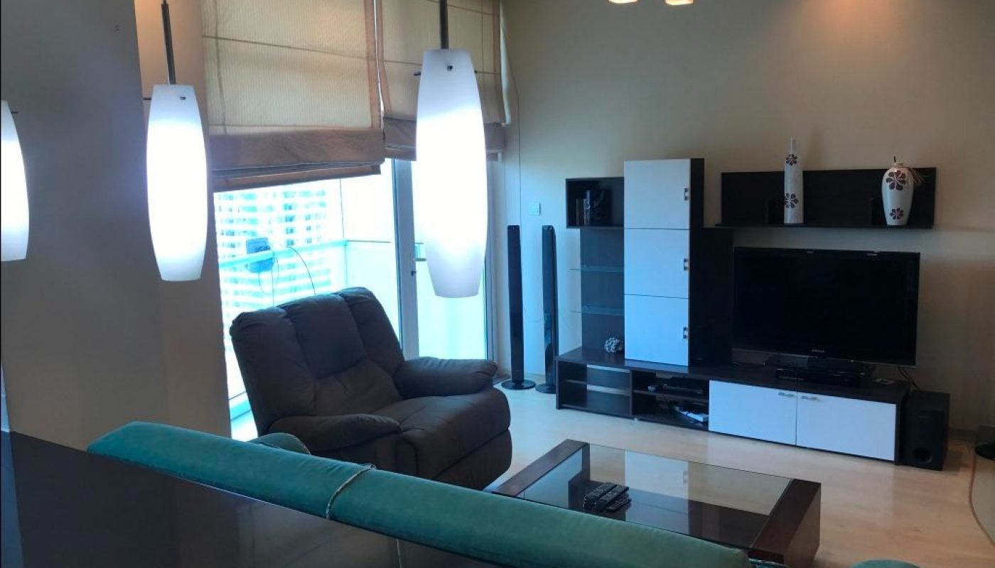 Modern living room with large windows, a recliner, entertainment center with TV, glass coffee table, and pendant lights.