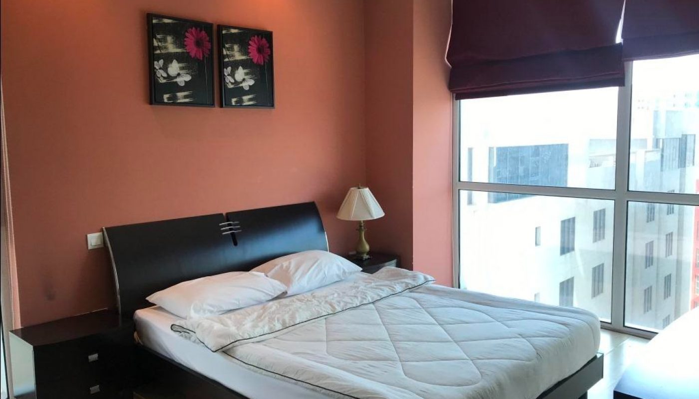 A neatly made double bed with white bedding is set against a coral-colored wall, featuring two framed pictures above the headboard and a window with purple blinds to the right.