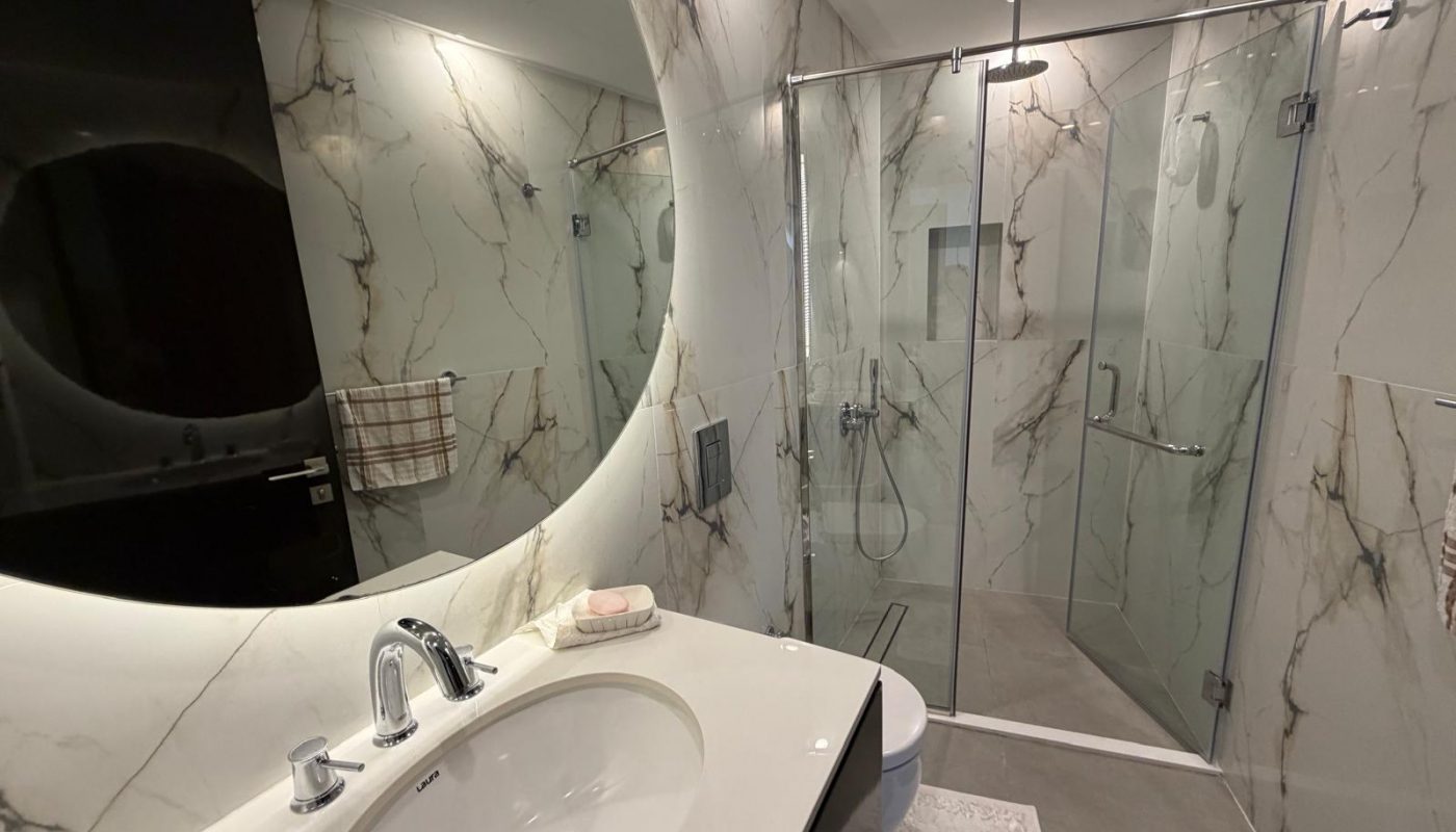 Modern bathroom with marble-patterned walls, a glass shower enclosure, a white sink with a chrome faucet, and a towel hanging on a rack.