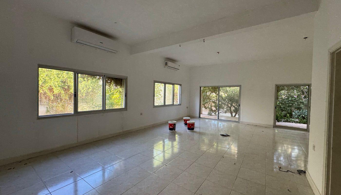 Spacious empty tiled room with large windows, two air conditioning units, three red paint buckets, and outdoor greenery visible through the windows and glass doors.