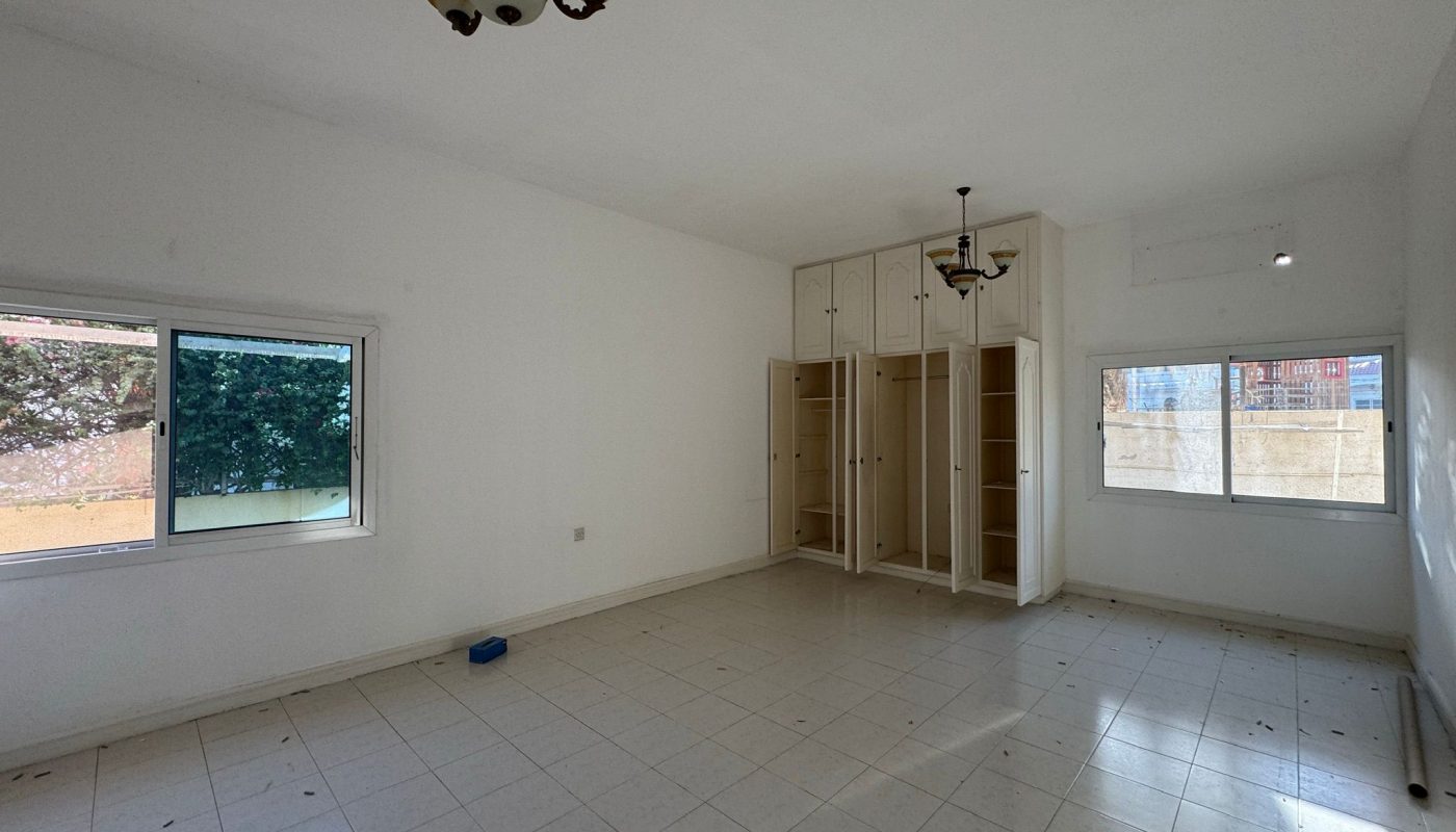 Empty room with white tiled floor, built-in open wardrobes, two windows, and a ceiling light fixture. A small blue object is on the floor near the corner.