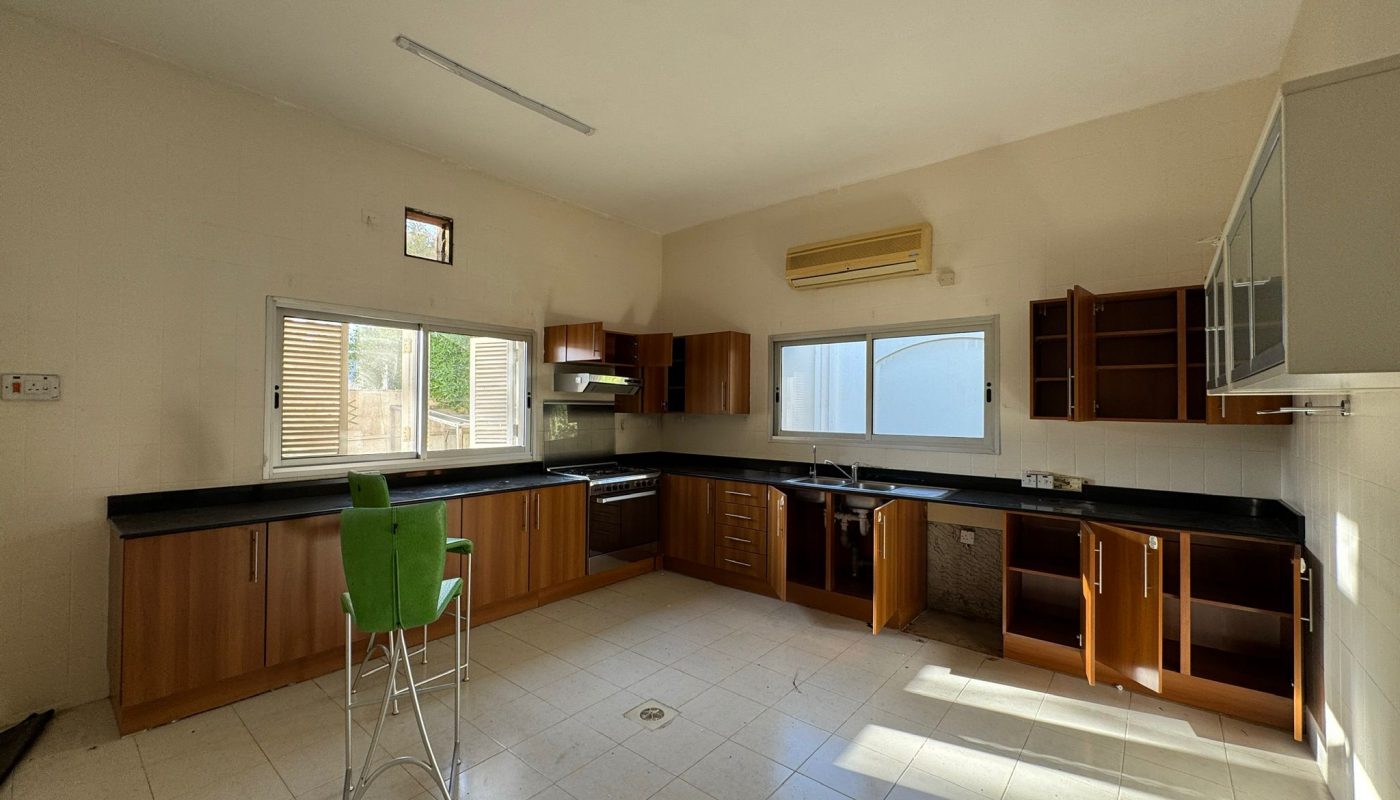 Spacious kitchen with wooden cabinets, black countertops, some open cabinet doors, a stove, sink, two green chairs, tiled floor, and large windows letting in sunlight.