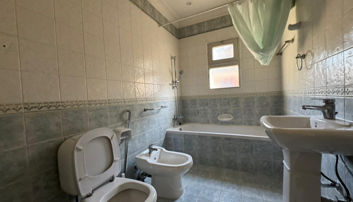 A bathroom with a toilet, bidet, bathtub with shower, sink, tiled walls and floor, window, and a green shower curtain draped to one side.