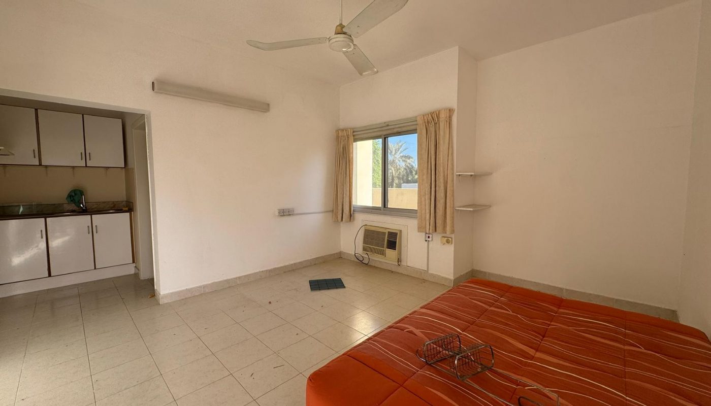 A sparsely furnished room with a bed, ceiling fan, window air conditioner, tiled floor, and a small kitchenette in the corner.