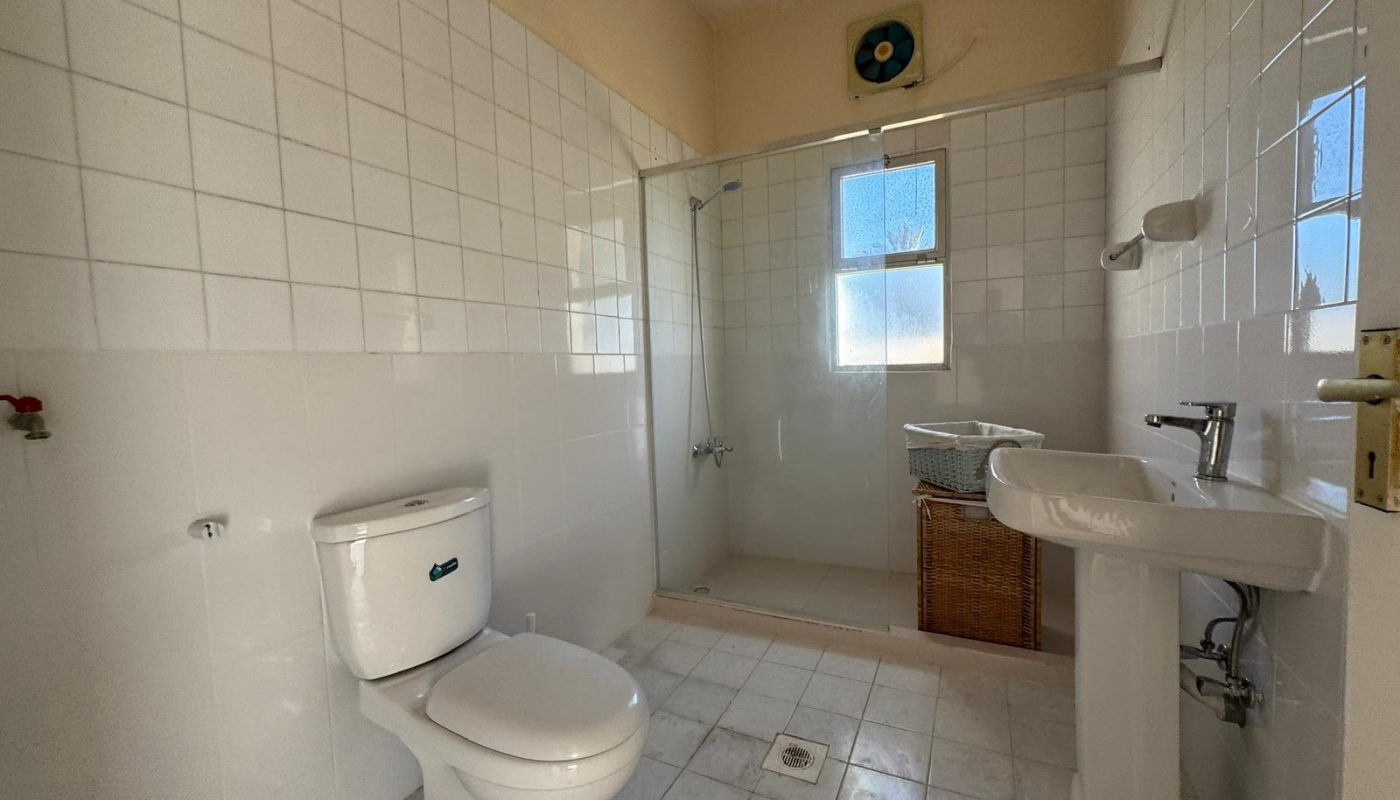A bathroom with a toilet, pedestal sink, glass-enclosed shower, and two windows; white tiles cover the walls and floor. A wicker basket sits near the shower.