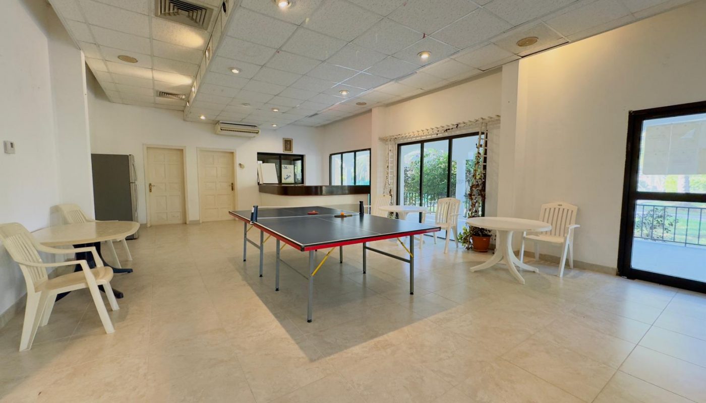Spacious indoor room with a ping pong table in the center, surrounded by white plastic chairs and tables, large windows, and tiled floor.