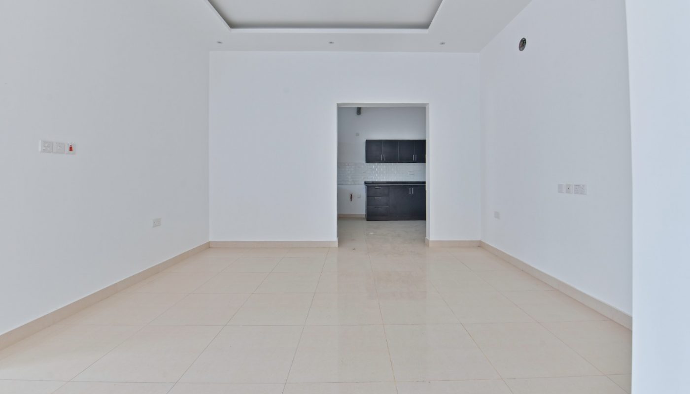 Empty, unfurnished room with light-colored tile flooring and white walls, featuring a recessed ceiling and a doorway leading to a kitchen area with dark cabinets.