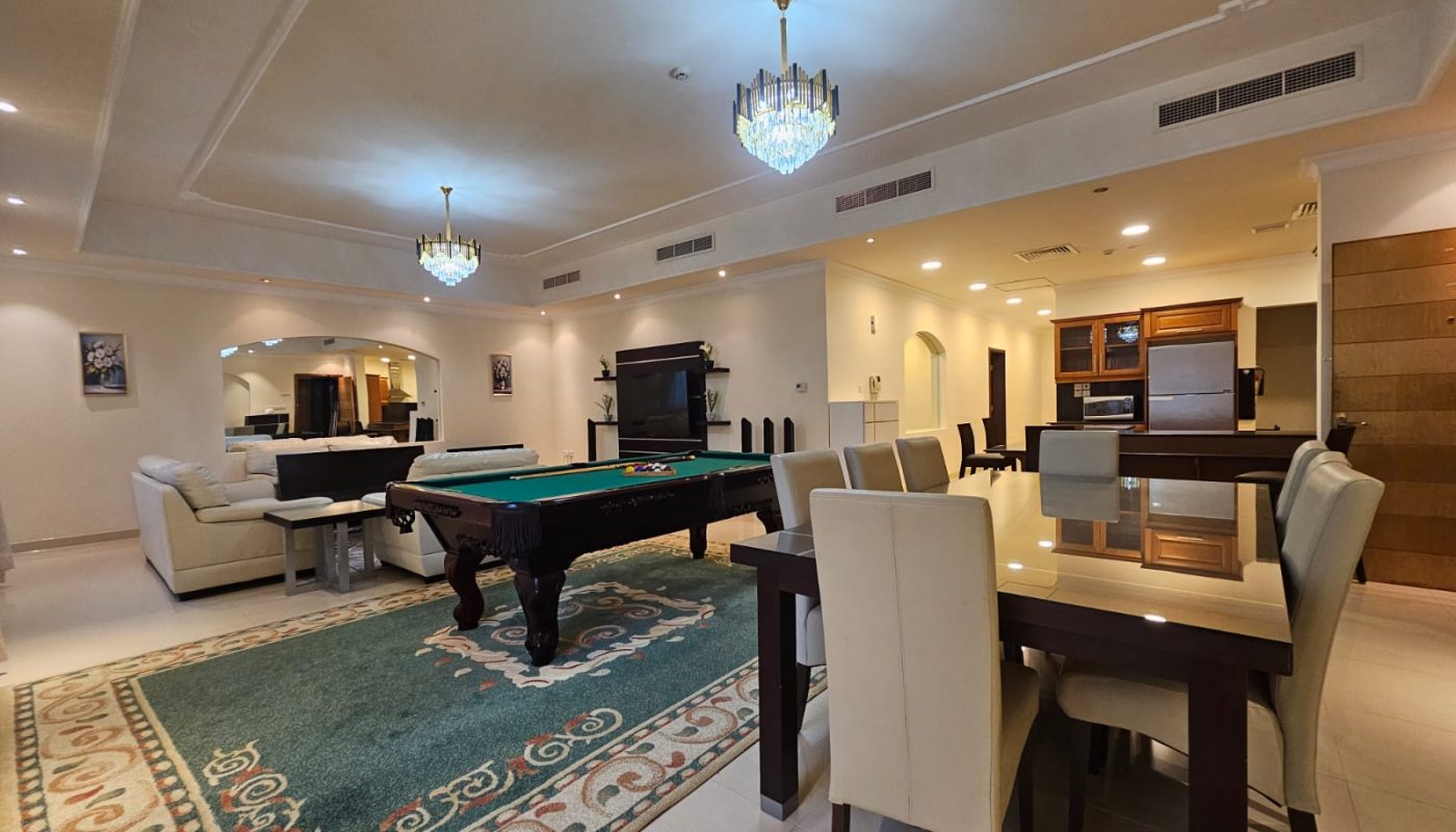 Spacious living area with a pool table, dining table, sofas, and a modern kitchen, illuminated by chandeliers and recessed lighting.
