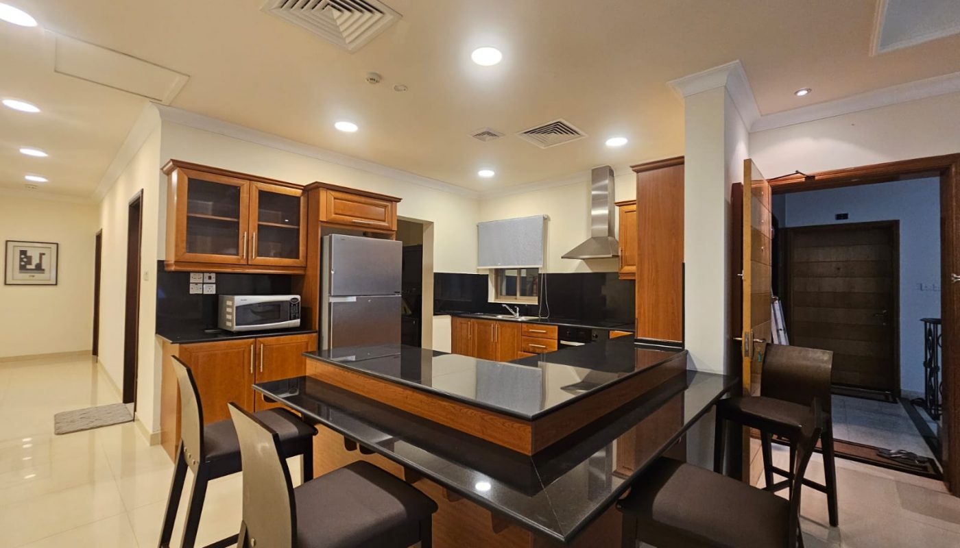 Modern kitchen with wooden cabinets, black countertops, stainless steel appliances, an island with seating for four, and bright ceiling lights. A hallway and door are visible in the background.