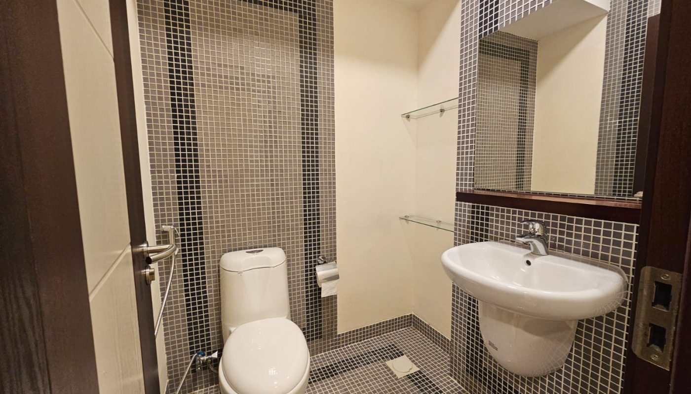 A small bathroom with a toilet, wall-mounted sink, large mirror, and tiled walls and floor in a grid pattern. Glass shelves are mounted above the sink.