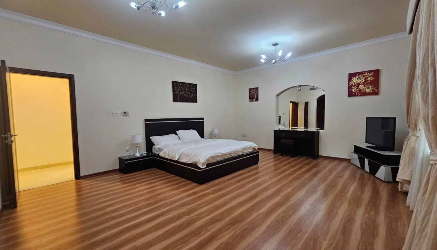 A spacious bedroom with a double bed, bedside tables, dressing table with mirror, wall-mounted TV, and wooden flooring under bright ceiling lights.