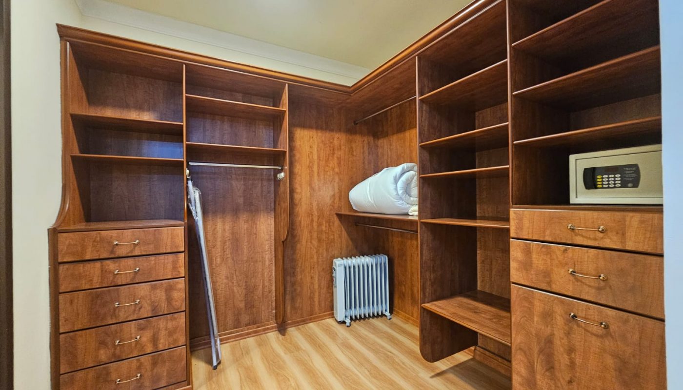 Walk-in closet with wooden shelves, drawers, hanging rods, a wall safe, an ironing board, radiator, and folded bedding on a shelf.