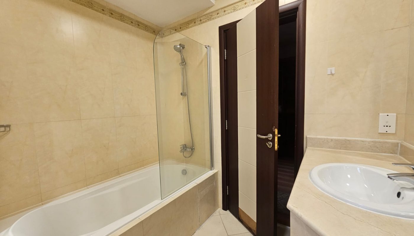 A bathroom with a bathtub and shower, a glass shower screen, a sink with a countertop, beige tiled walls, and an open wooden door.