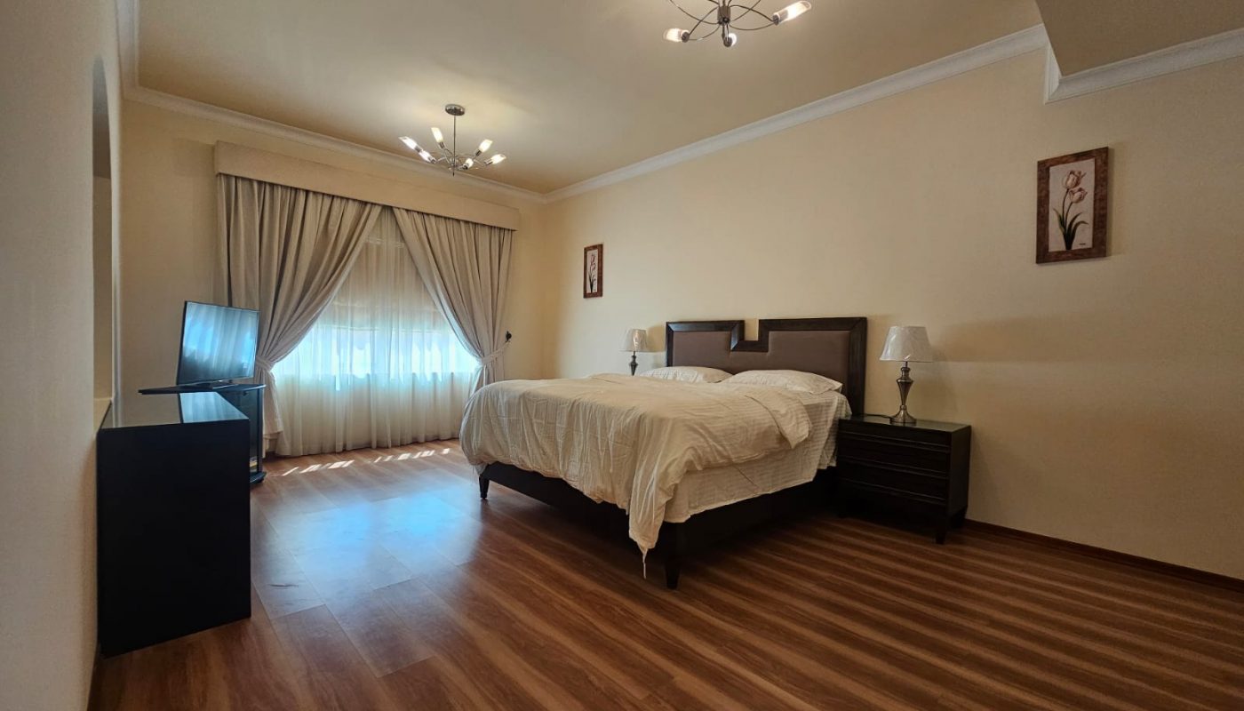 A spacious bedroom with a large bed, two nightstands, a TV on a dresser, wooden floors, and a window with beige curtains letting in natural light.