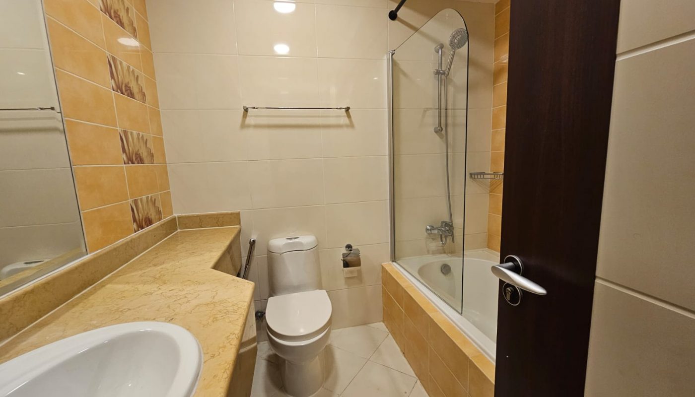 Modern bathroom with beige tiles, a countertop sink, a toilet, a towel rack, and a bathtub with a glass shower screen and wall-mounted showerhead. Door partially open.