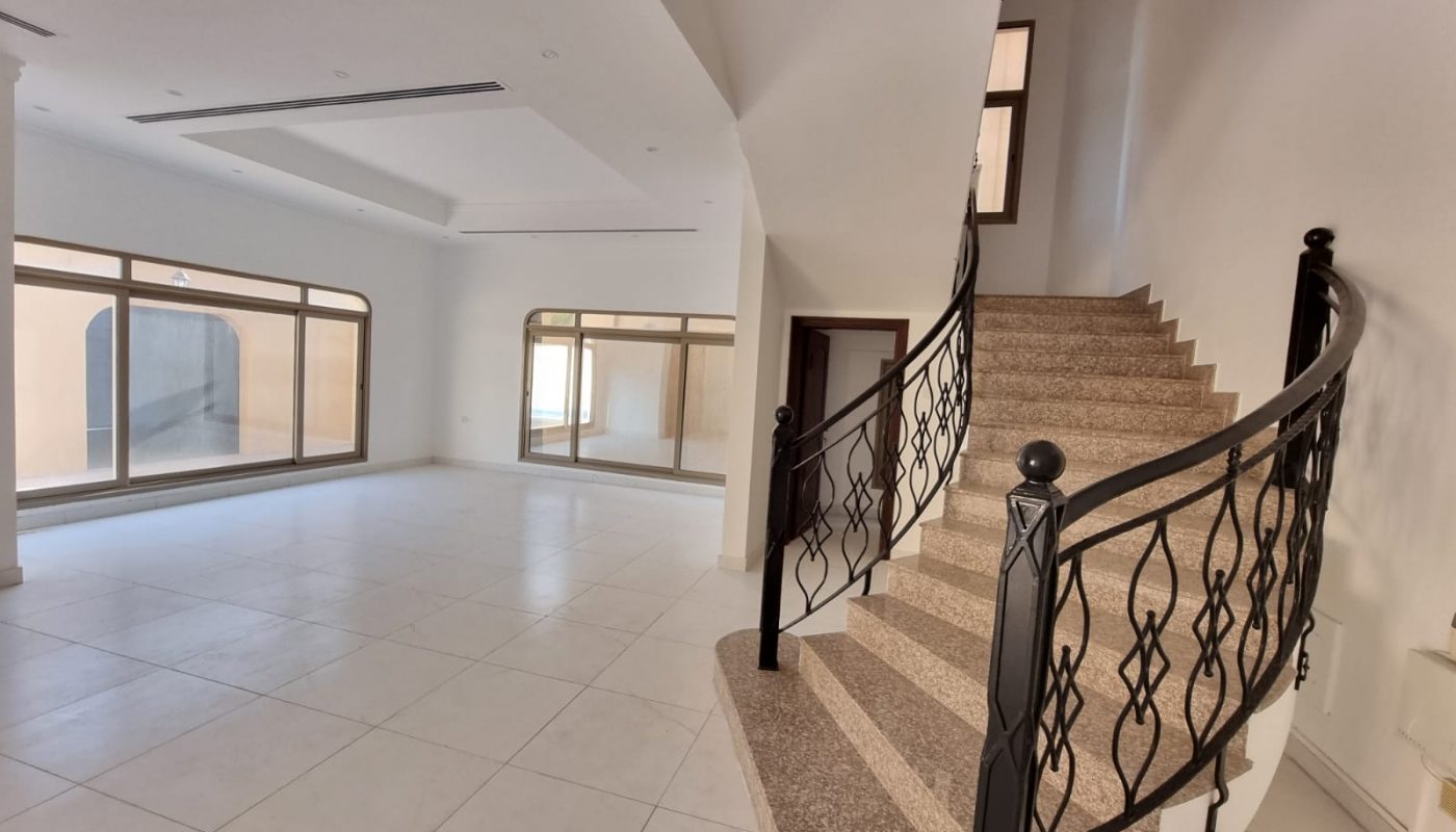 Spacious empty room with white tiled floor, large windows, and a carpeted staircase with a black wrought iron railing.