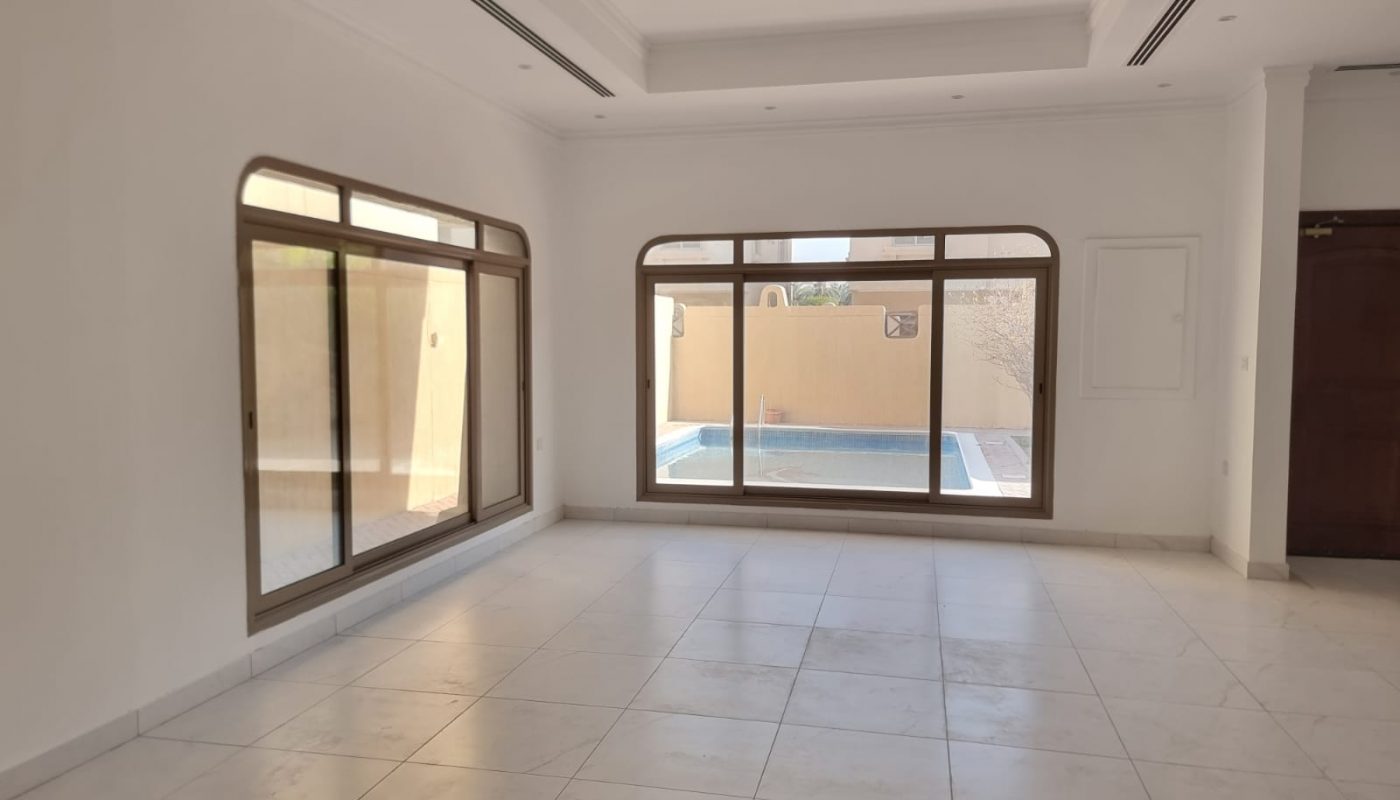 Spacious empty room with large windows, tiled floors, white walls, and a view of a swimming pool and courtyard outside.