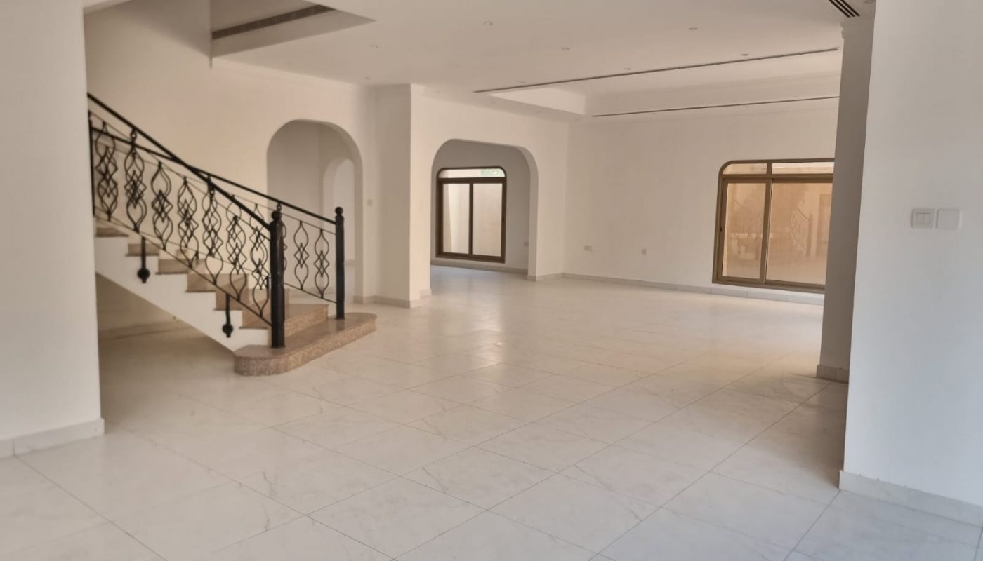 Spacious empty room with white tiled floors, large windows, arched doorways, and a staircase with a decorative black railing.