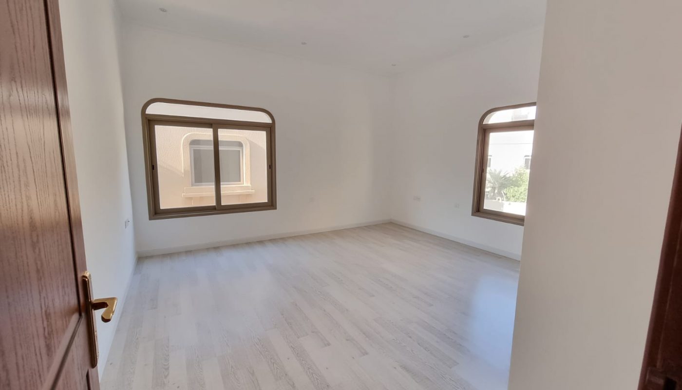 Empty, unfurnished room with light wood flooring, white walls, two large windows, and a wooden door partially open. Natural light is coming through the windows.