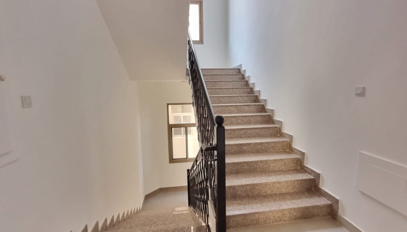Indoor staircase with brown marble steps, black decorative railing, and white walls, leading up to a window with natural light coming through.
