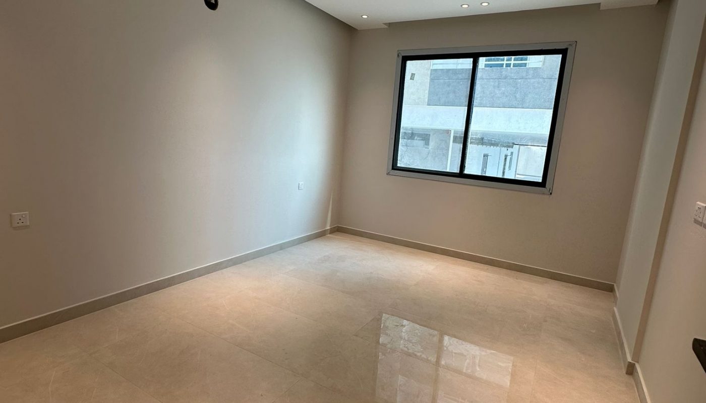 Empty, unfurnished room with beige tiled floor, white walls, recessed ceiling lights, and a large window showing a view of another building.