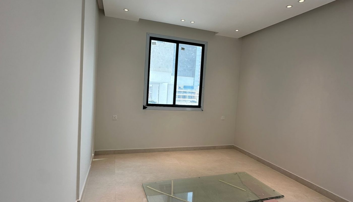 Empty room with beige tiled floor, white walls, recessed ceiling lights, a single window, and a large glass sheet lying on the floor.