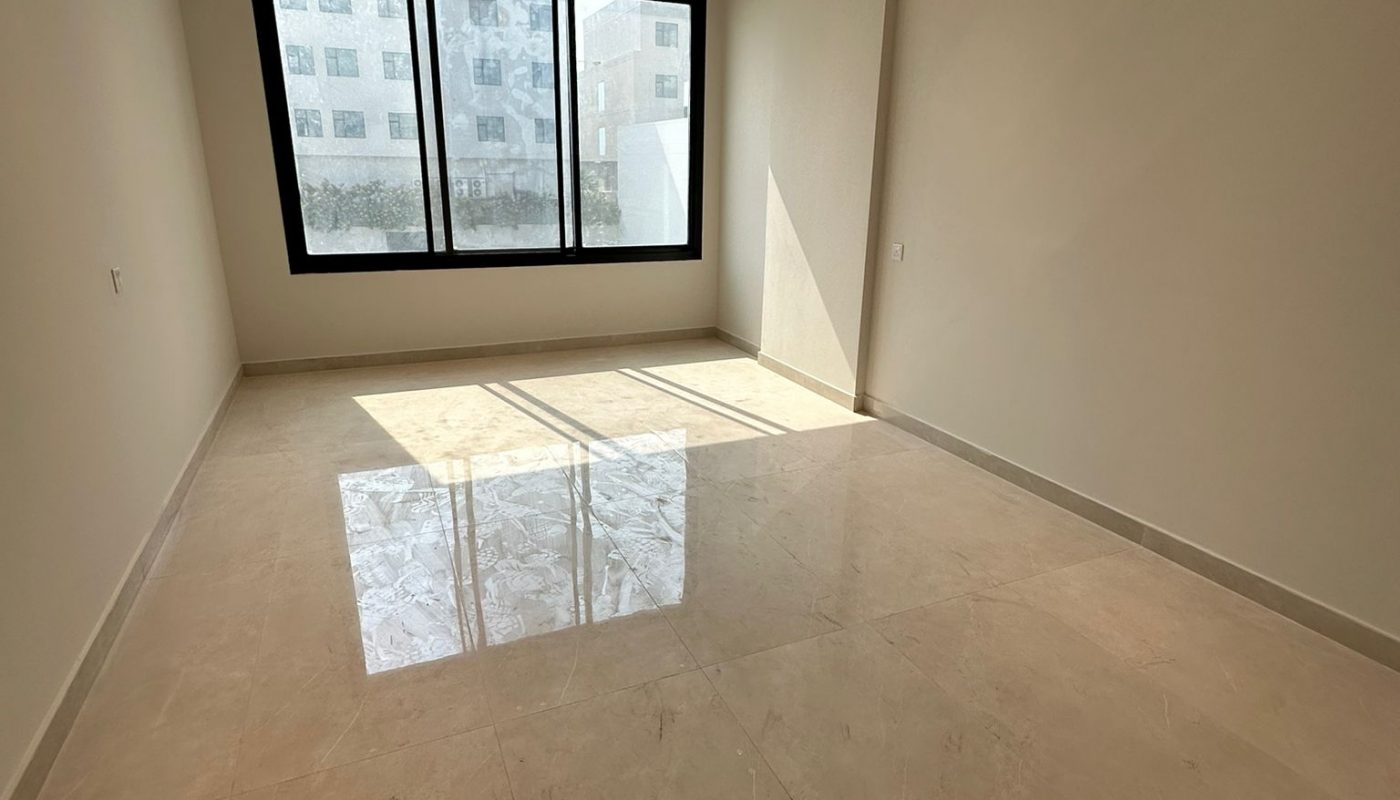 Empty room with large window, beige tiled floor, white walls, and ceiling lights. Sunlight is streaming in, creating reflections on the floor.