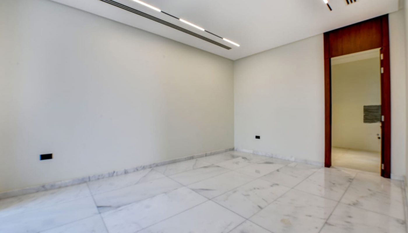 Spacious empty room with white marble tile flooring, white walls, modern ceiling lights, and a wooden door slightly open to an adjacent area.